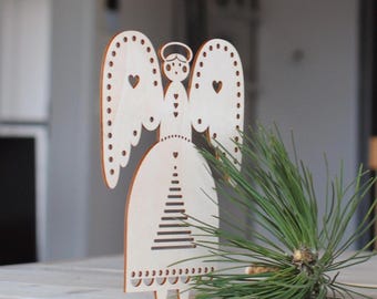 Op de afbeelding: Een houten engelenbeeldje met een halo, hartvormige uitsparingen en een jurk met een boomontwerp. De engel staat op een lichtgekleurd oppervlak, met een takje groene dennennaalden op de voorgrond.