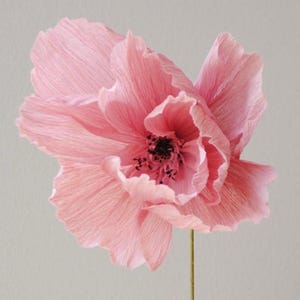 Puede incluir: Una flor de papel rosa claro hecha a mano, con un centro oscuro y un tallo verde fino. Los p&eacute;talos de la flor son rizados y texturizados, creando una apariencia delicada y realista. La flor est&aacute; sobre un fondo gris claro.
