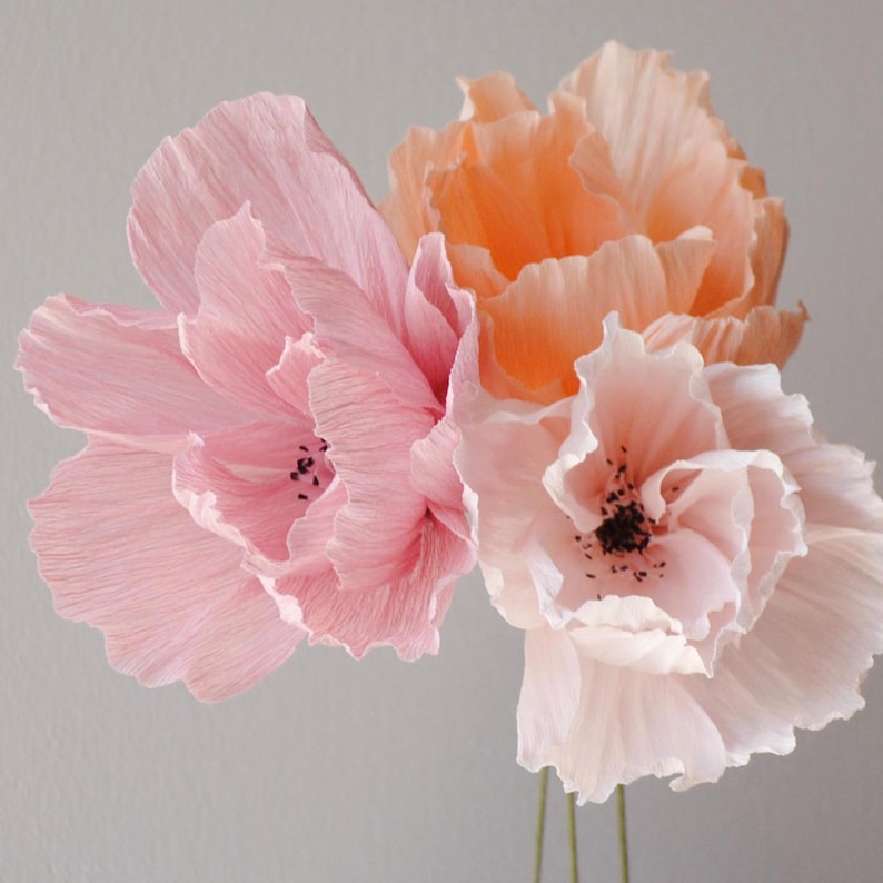 Puede incluir: Tres flores de papel en tonos rosa y melocot&oacute;n. Las flores tienen p&eacute;talos texturizados y centros oscuros. Los tallos son finos y verdes. Las flores est&aacute;n dispuestas sobre un fondo gris claro.