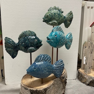 Könnte beinhalten: Vier Keramik-Fischfiguren in Blautönen und Grün sind auf Holzstäben montiert und auf einem Holzscheit-Sockel angeordnet. Die Fische sind alle in verschiedene Richtungen ausgerichtet und haben unterschiedliche Designs.