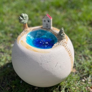 Könnte beinhalten: Eine kugelförmige Keramikskulptur mit einer Miniaturlandschaft. Sie zeigt ein blaues Wasserelement, ein kleines Haus mit rotem Dach und einen Baum. Die Skulptur ist hauptsächlich cremefarben mit blauen, braunen und grünen Akzenten.
