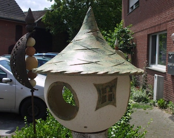 Op de afbeelding: Een keramisch vogelhuisje met een groen dak en witte muren. Het vogelhuisje heeft een klein raam en een ronde opening. Het vogelhuisje staat op een houten paal.