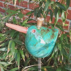 May include: A teal ceramic bird garden stake with a long, orange beak and a small crown. The bird has brown accents and a red eye. It is set against a backdrop of green bamboo and a red brick wall.