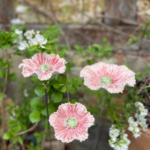 Könnte beinhalten: Drei Gartenstecker aus Keramik in Form von Blumen mit rosa Blütenblättern und grünen Zentren. Die Stecker werden zwischen grünem Laub und weißen Blüten gezeigt, mit einer verschwommenen Backsteinmauer im Hintergrund. Die Blumen haben ein strukturiertes Aussehen.
