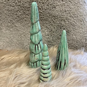 Könnte beinhalten: Drei Keramik-Weihnachtsbaumfiguren in verschiedenen Größen. Die Bäume sind helltürkis mit dunklen Akzenten und stehen auf einer flauschigen, hellen Oberfläche. Der größte Baum ist etwa 15 cm hoch.