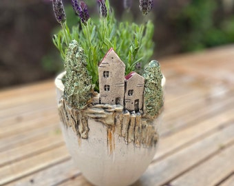 Może przedstawiać: Ceramiczna donica z fantazyjną sceną wioski, z miniaturowymi domami z czerwonymi dachami i teksturowanymi drzewami. Donica jest wypełniona roślinami lawendy z fioletowymi kwiatami, tworząc uroczy, dekoracyjny element.