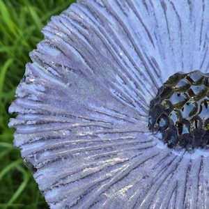 May include: A circular ceramic sculpture featuring a textured, light purple surface with radiating grooves that extend from the center. The center of the piece is a dark, glossy, honeycomb-like structure with deep, recessed, and reflective black cells.