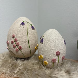 May include: Two decorative egg-shaped ornaments with a textured, off-white surface. Each egg features painted floral designs, including purple bellflowers and pink and yellow spiral-shaped flowers. The eggs are placed on a fluffy, light-coloured surface.