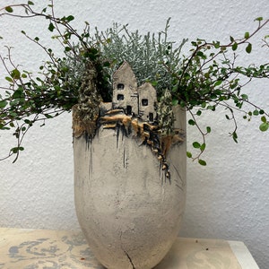 Può includere: Un vaso di ceramica a forma di scogliera con un piccolo villaggio in cima. Il vaso è dipinto in tonalità di bianco, grigio e oro. Il vaso è pieno di fogliame verde e argentato.