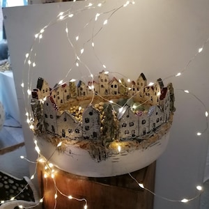 Op de afbeelding: Een witte keramische kom met een gedetailleerde miniatuur dorpsscène rond de rand. De huizen hebben rode, zwarte en witte accenten. Twinkelende lichtslingers zijn om de kom gedrapeerd, wat een warme gloed creëert.