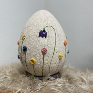 Op de afbeelding: Een groot, eivormig decoratief object met een getextureerd, gebroken witte oppervlak. Het heeft kleurrijke, verhoogde bloemaccenten in paars, geel, roze en oranje, met groene stelen. Het object staat op een zacht, lichtgekleurd oppervlak.