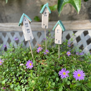 Op de afbeelding: Drie miniatuurhuisjes met groene daken en witte muren zijn in een groepje gerangschikt op een bed van paarse bloemen en groen gebladerte.