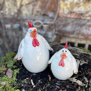 Può includere: Due statuette di galline in ceramica. Le galline sono bianche con creste e bargigli rossi e becchi arancioni. Hanno piccole ali e sono sedute su un letto di muschio e terra. Le statuette hanno un aspetto rustico e fatto a mano.