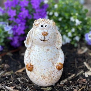 Puede incluir: Figura de oveja de cerámica con cuerpo blanco moteado y detalles marrones en las orejas, la nariz y las patas. La oveja tiene una expresión sonriente y está colocada sobre un fondo de flores moradas y blancas.