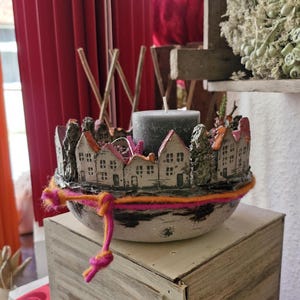 Könnte beinhalten: Ein dekorativer Kerzenhalter mit einem Miniatur-Dorfdesign. Der Halter ist hellgrau, mit kleinen weißen Häusern und orangefarbenen und rosa Dächern. Eine graue Kerze befindet sich in der Mitte, und bunte Wolle ist um die Basis gewickelt.