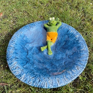 Könnte beinhalten: Eine blaue Vogeltränke aus Keramik mit strukturierter Oberfläche. Eine grüne Froschfigur mit orangefarbenen Shorts liegt in der Mitte. Die Vogeltränke steht auf grünem Gras.