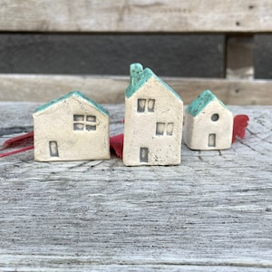 Könnte beinhalten: Drei kleine Keramikhäuser mit türkisfarbenen Dächern. Die Häuser sind weiß mit schwarzen Fenstern und Türen. Die Häuser stehen auf einer Holzoberfläche.