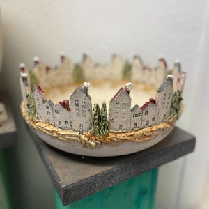 Könnte beinhalten: Eine weiße Keramikschale mit einem dekorativen Design einer kleinen Stadt mit Häusern und Bäumen am Rand. Die Schale steht auf einem Holzständer.