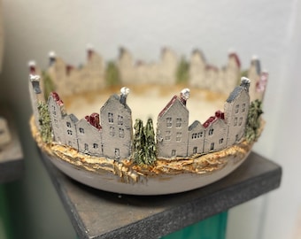 Może przedstawiać: Biała ceramiczna miska z dekoracyjnym wzorem przedstawiającym małe miasteczko z domami i drzewami wokół krawędzi. Miska stoi na drewnianym stojaku.