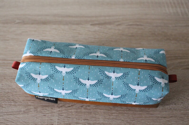 May include: A blue and white zippered pouch with a pattern of white cranes flying against a blue background. The pouch has a brown leather bottom and a red zipper pull.