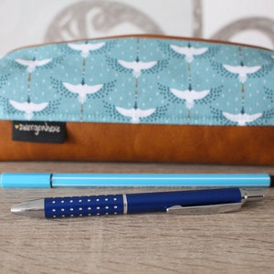 May include: A brown leather pencil case with a blue and white fabric lining featuring a pattern of white birds. The case is closed and has a small white label with the text "zwengerhohe" on it. Two pens are lying on a wooden surface in front of the case.