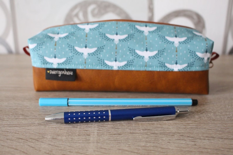 May include: A brown leather pencil case with a blue and white fabric lining featuring a pattern of white birds. The case is closed and has a small white label with the text "zwengerhohe" on it. Two pens are lying on a wooden surface in front of the case.