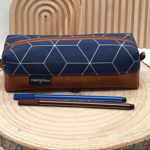 Könnte beinhalten: Rechteckiges Federmäppchen mit marineblauem geometrischen Muster und braunem Lederboden. Das Etui hat einen Reißverschluss und ein kleines Etikett mit Text. Zwei Stifte liegen vor dem Etui.