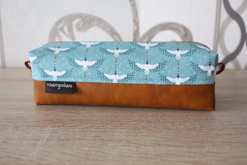 May include: A brown leather pencil case with a blue fabric top featuring a white crane pattern. The fabric has a gold and blue dot pattern. The case has a small label that says "zwergenhexe".