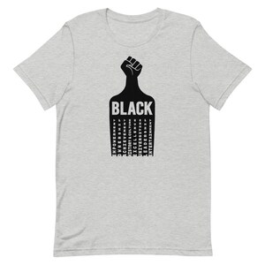 K&ouml;nnte beinhalten: Ein graues T-Shirt mit einer schwarzen Faust, die in die Luft gehoben wird. Das Wort "BLACK" ist in schwarzen Buchstaben unter der Faust geschrieben. Die W&ouml;rter "EMPOWERMENT", "OWNERSHIP", "ACHIEVEMENT", "HISTORY & CULTURE", "UNIVERSITIES", "EXCELLENCE", "LEADERS", "ENTERTAINMENT" sind in einer Spalte unter dem Wort "BLACK" geschrieben.