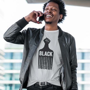 K&ouml;nnte beinhalten: Ein Mann tr&auml;gt ein graues T-Shirt mit einem schwarzen Faust-Motiv und dem Wort "BLACK" darauf gedruckt. Das Shirt zeigt auch eine Liste von W&ouml;rtern, die mit der schwarzen Kultur zusammenh&auml;ngen, darunter "Empowerment", "Achievement", "Diversity" und "Entertainment".