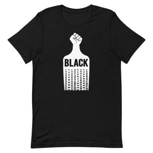 K&ouml;nnte beinhalten: Schwarzes T-Shirt mit einem erhobenen Faust-Motiv und dem Wort "BLACK" in wei&szlig;en Buchstaben. Das Wort "BLACK" ist umgeben von W&ouml;rtern, die die schwarze Erfahrung beschreiben, darunter "Empowerment", "Ownership", "Achievement", "History & Culture", "Excellence", "Universities", "Leaders" und "Entertainment".