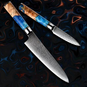 Pode incluir: Duas facas de cozinha em aço Damasco com cabos de madeira e resina azul. Os cabos apresentam um grão de madeira castanho claro e uma resina azul vibrante. As lâminas têm um padrão distinto.