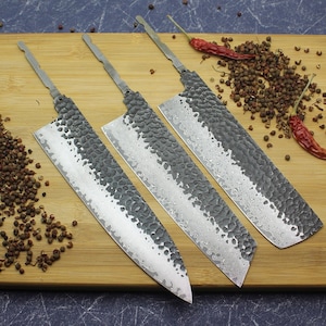 May include: Three kitchen knives with hammered steel blades and wooden handles. The blades are silver and black with a textured finish. The knives are arranged on a wooden cutting board.