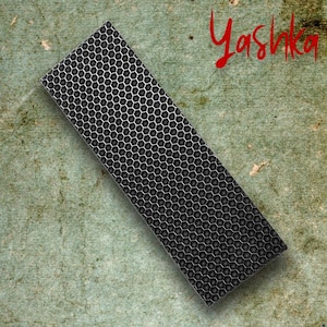 Może przedstawiać: Prostokątny metalowy przedmiot z wzorem plastra miodu, ustawiony pod kątem na teksturowanej zielonej powierzchni. Słowo "Yashka" jest napisane czerwoną czcionką w prawym górnym rogu. Przedmiot wydaje się być dekoracyjny lub funkcjonalny.