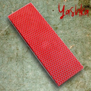 May include: A rectangular red box with a white honeycomb pattern. The word "Yashka" is written in red script in the upper right corner. The box is angled on a textured, green-toned surface.