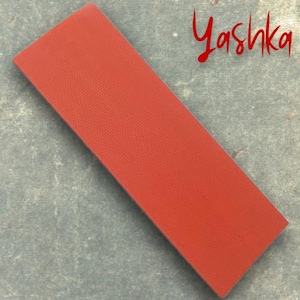 Może przedstawiać: Prostokątny, teksturowany, czerwony przedmiot ze słowem "Yashka" w czerwonym skrypcie. Obiekt jest ustawiony pod kątem na szarej powierzchni. Czerwony obiekt wydaje się być wykonany z elastycznego materiału.