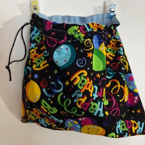 May include: A black drawstring bag with a colorful "Happy Birthday" print. The design includes balloons, confetti, and planets in various colors. The bag is lined with light blue fabric.