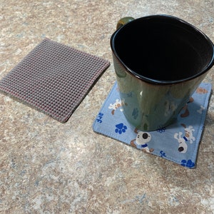 May include: Two square coasters, one gray and one blue with a white paw print and dog design. A dark green mug sits on the blue coaster.