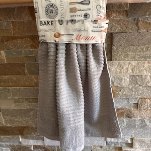 May include: A hanging kitchen towel featuring a grey ribbed fabric attached to a cream-colored fabric top with a kitchen-themed print. The top section displays illustrations of whisks, spoons, and text including BAKE, SALADE, and MENU.