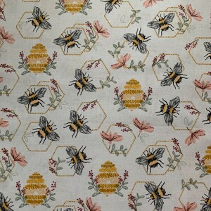 May include: A light-colored fabric featuring a repeating pattern of embroidered bumblebees, golden yellow beehives, and hexagonal outlines. The design includes small pink flowers and green sprigs interspersed throughout the repeating motif.