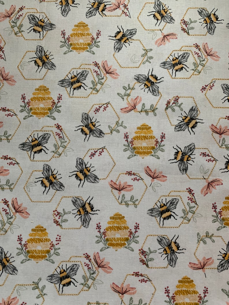 May include: A light-colored fabric featuring a repeating pattern of embroidered bumblebees, golden yellow beehives, and hexagonal outlines. The design includes small pink flowers and green sprigs interspersed throughout the repeating motif.