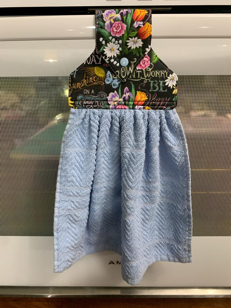 May include: A hanging kitchen towel with a light blue textured fabric and a black fabric top featuring a colorful floral print, a light blue snap button, and the text "Sunshine on a Cloudy Day" and "Don't Worry Be Happy" in white script.