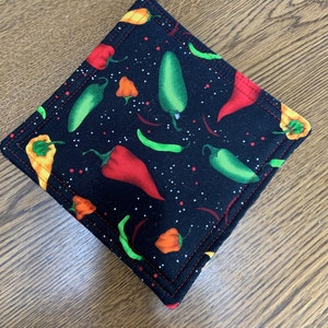 May include: A black fabric hot pad with a red stitched border. The fabric has a pattern of green and red peppers on a black background with white speckles.