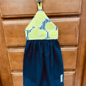 May include: A blue kitchen towel with a lemon print hanging on a cabinet door. The towel has a loop for hanging and a blue and white striped fabric panel with lemons.