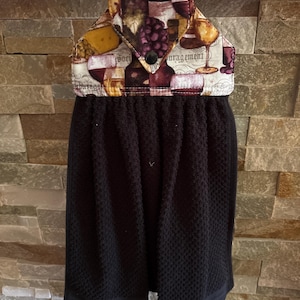 May include: A black hanging kitchen towel with a fabric top featuring a wine and grape print. The top section has a pointed flap secured by a black button and displays illustrations of wine glasses, bottles, and grapes with the text "Soci d'ourage".