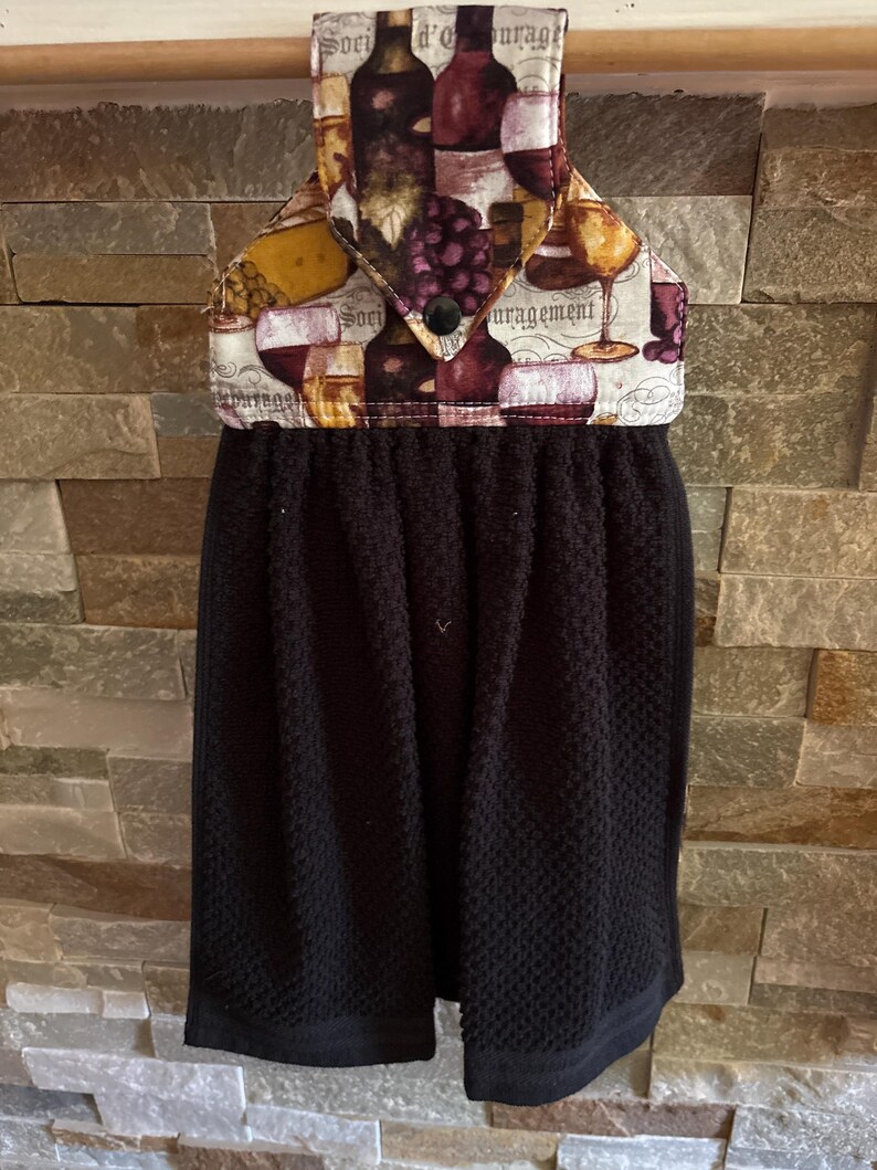 May include: A black hanging kitchen towel with a fabric top featuring a wine and grape print. The top section has a pointed flap secured by a black button and displays illustrations of wine glasses, bottles, and grapes with the text "Soci d'ourage".