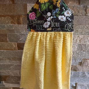 May include: A hanging kitchen towel featuring a bright yellow, textured fabric attached to a black fabric top with a floral pattern of sunflowers and daisies. The top section includes a white snap button and the text "DON'T WORRY YOU CAN GROW".