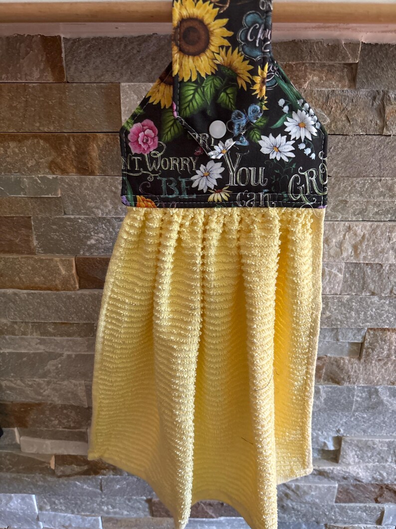 May include: A hanging kitchen towel featuring a bright yellow, textured fabric attached to a black fabric top with a floral pattern of sunflowers and daisies. The top section includes a white snap button and the text "DON'T WORRY YOU CAN GROW".