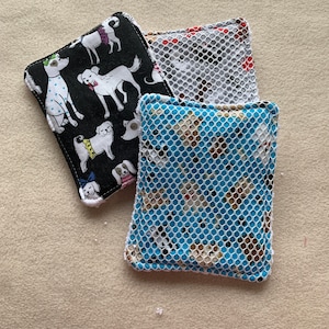 May include: Three reusable dishcloths with a white mesh backing. The front of the cloths feature a black and white dog print, a white and gray dog print, and a blue and white dog print.
