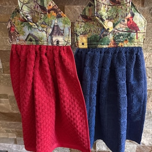 May include: Two hanging kitchen towels with a birdhouse and garden print on the top fabric panels. One towel is solid red with a textured weave, and the other is solid navy blue with a textured weave. Each towel features a snap button closure.